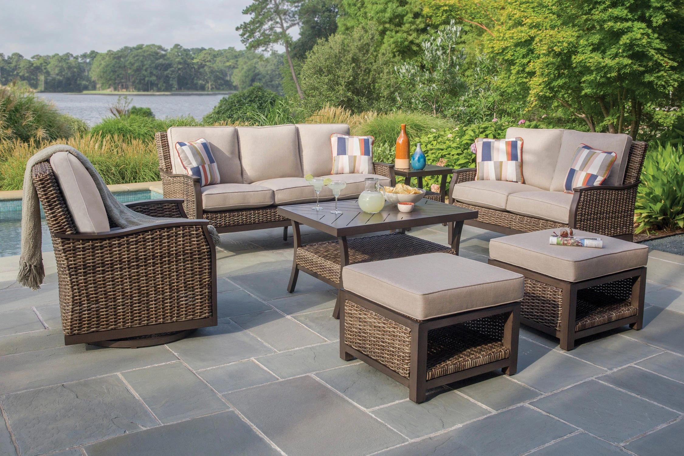 Outdoor patio furniture set with sofa, chairs, ottoman, and table on a stone terrace by landscaped greenery and water